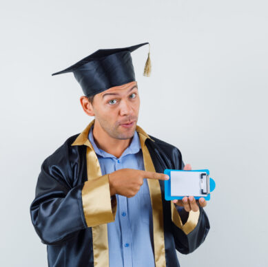 young male in graduation gown,cap pointing to mini clipboard and looking focused , front view.